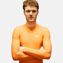 Ciclista con maillot de manga larga naranja Pas Normal Studios Mechanism, ajuste aerodinámico para ciclismo de ruta.